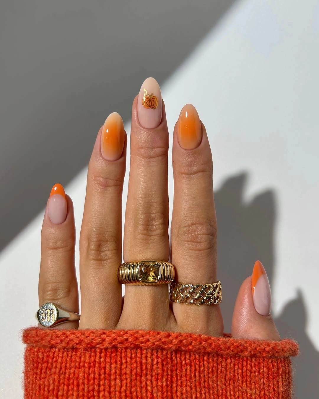 Short oval pale nails with warm orange French tips and a gold accent
