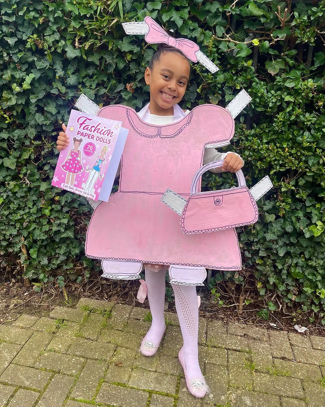 Child in pink paper-doll fashion Halloween costume with cardboard dress bow printed handbag and pink boots