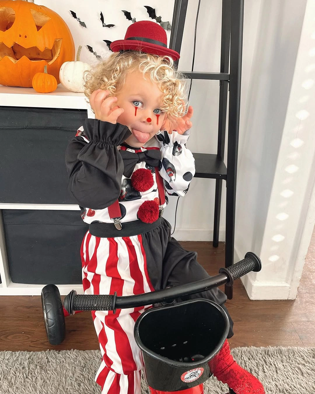 Child in red-and-white striped creepy clown Halloween costume with black puff sleeves black hat red bow and prop
