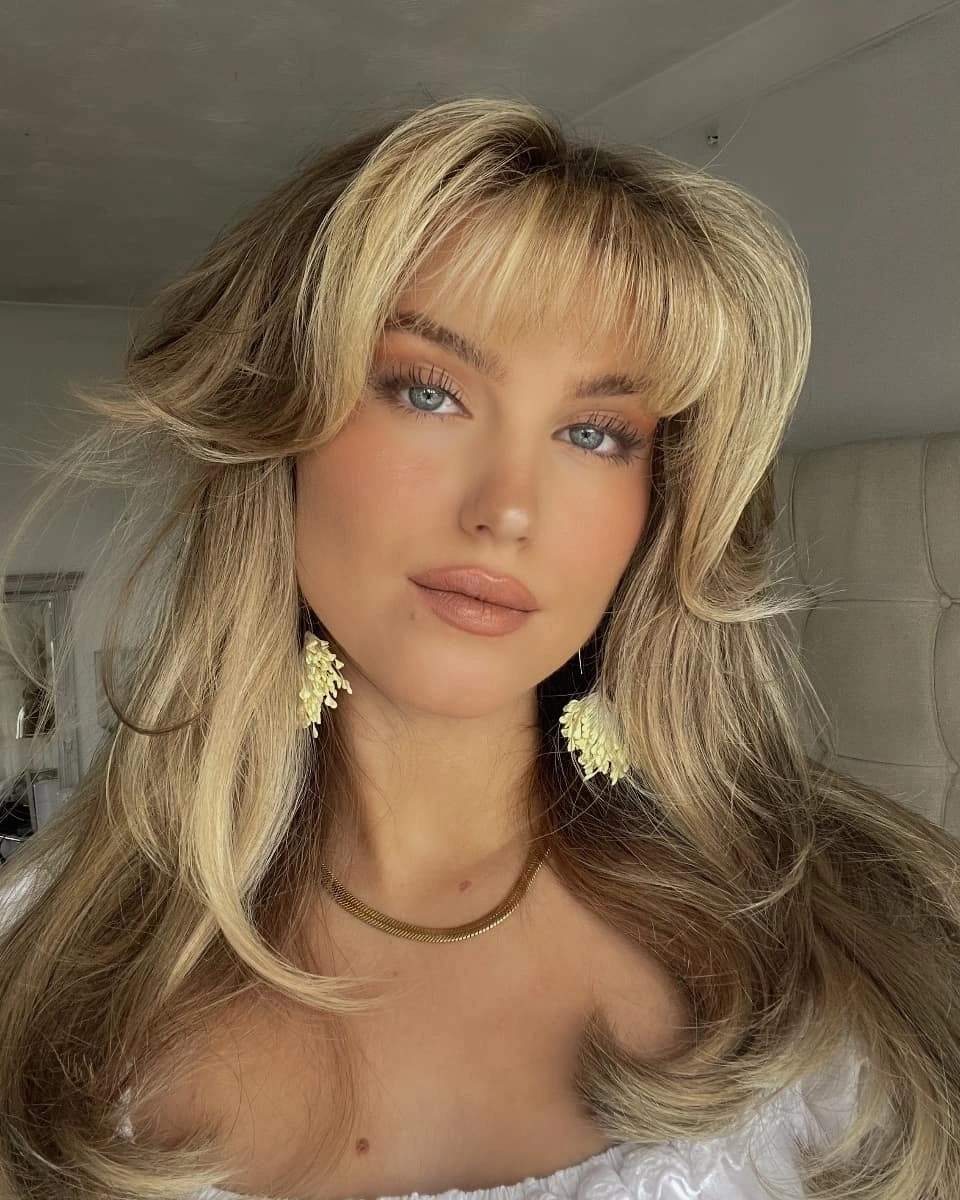 Long blonde layers with curtain fringe, soft root depth, and large face-framing waves