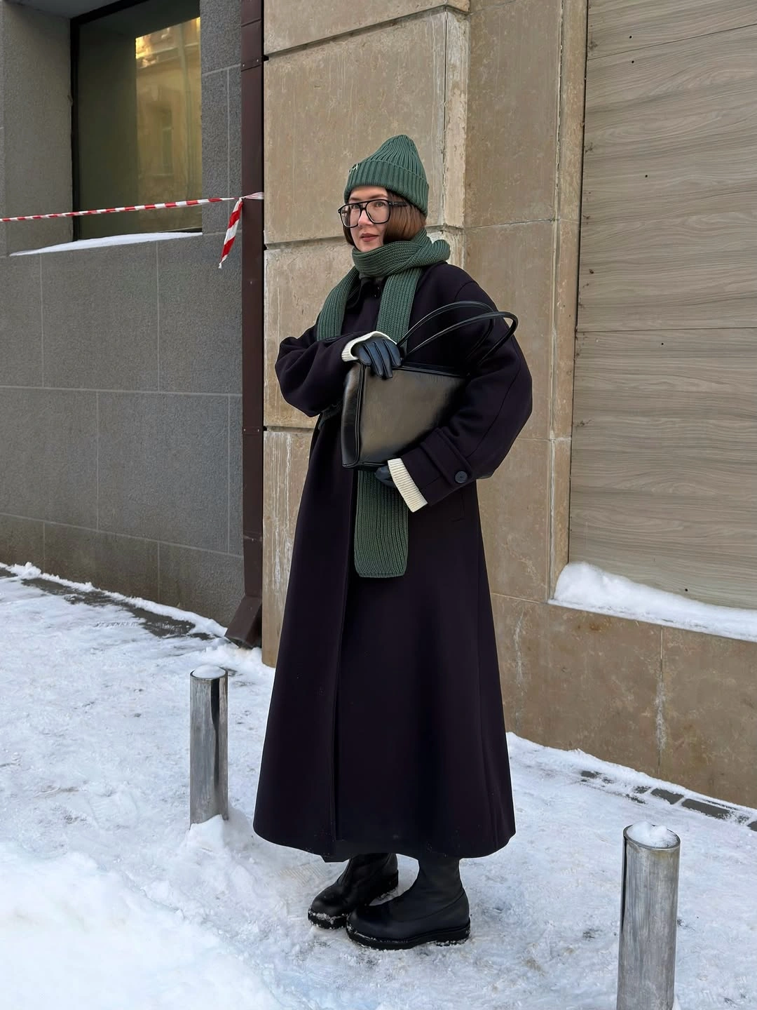 Black long coat green beanie green scarf dark trousers black boots and bag