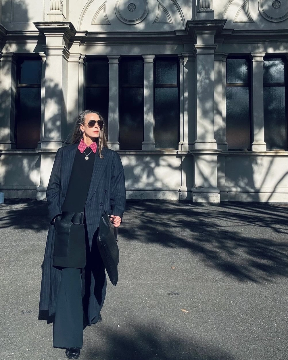 Long dark coat small pink neck scarf dark trousers sunglasses and black tote