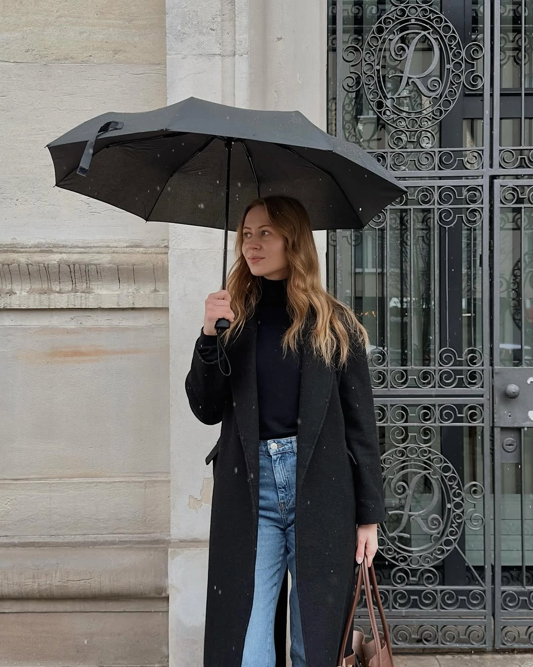Black long coat black top blue straight jeans black boots umbrella and handbag