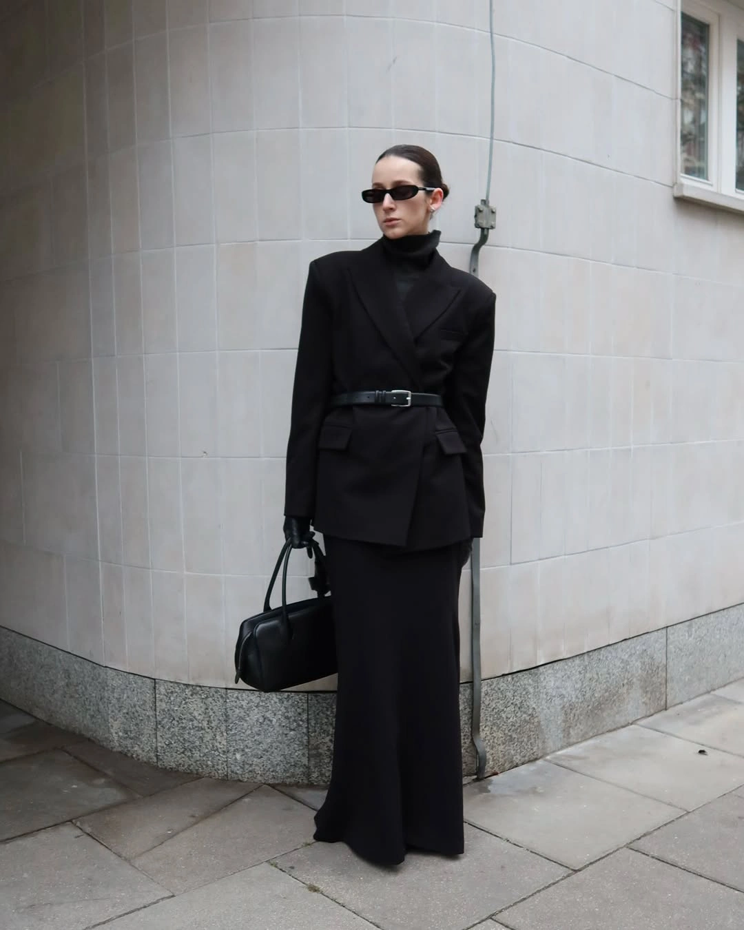 Black belted high neck jacket wide black trousers sunglasses and dark handbag