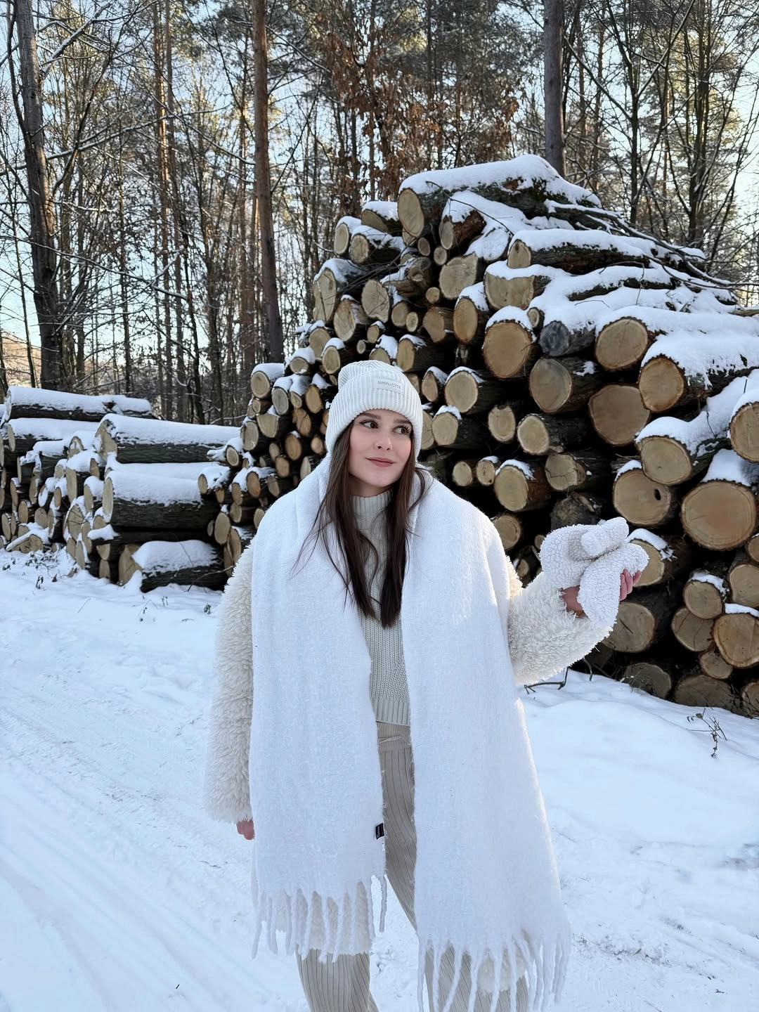 Woman in white sweater scarf hat and pale pants by stacked logs