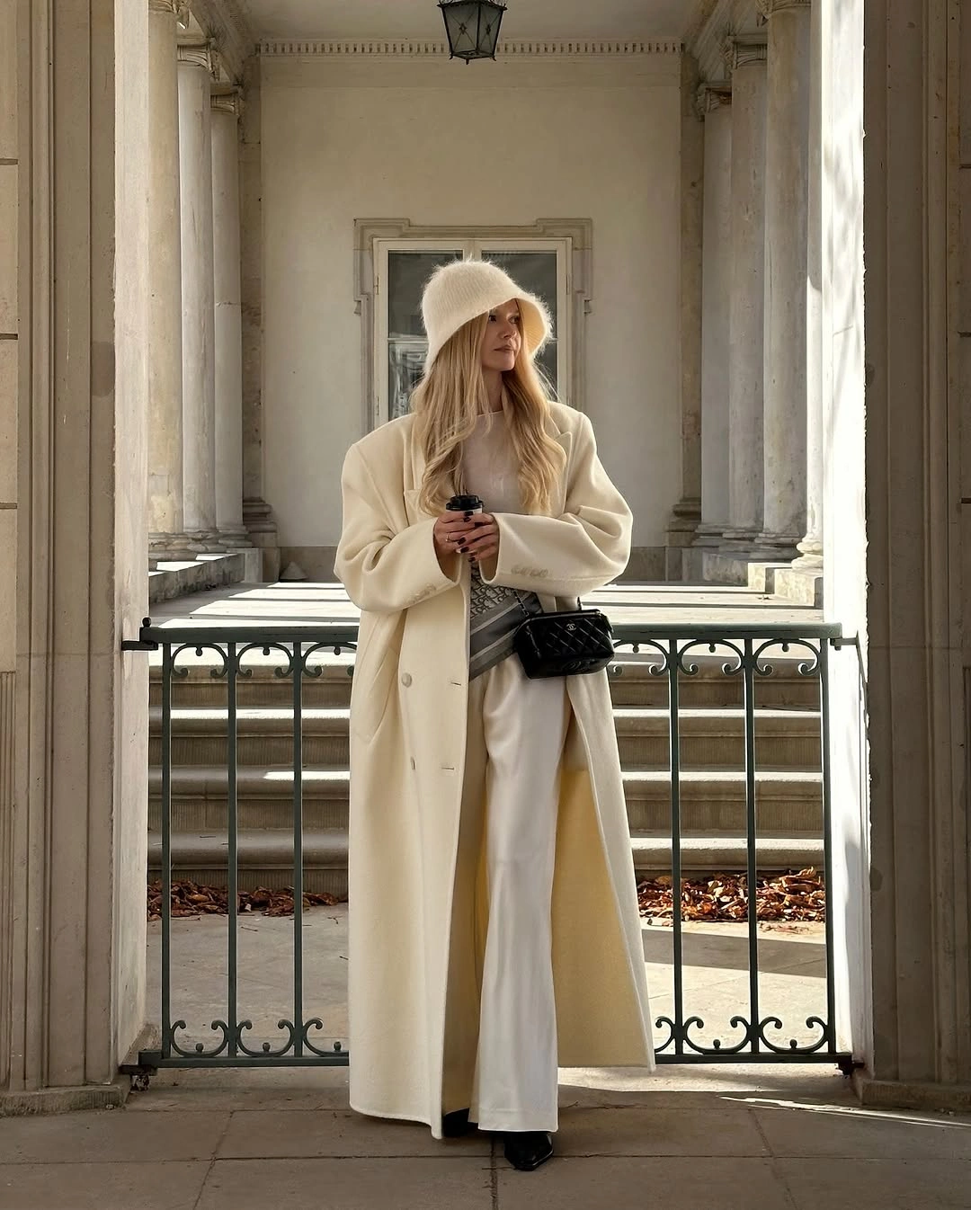 Woman in cream coat bucket hat white trousers and black bag