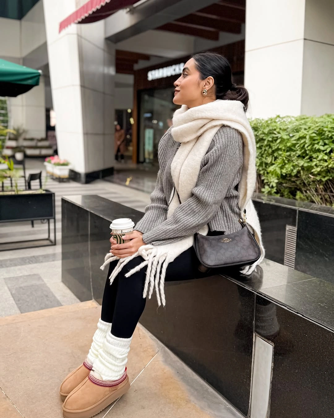 Woman in gray knit sweater cream scarf black leggings and tan boots