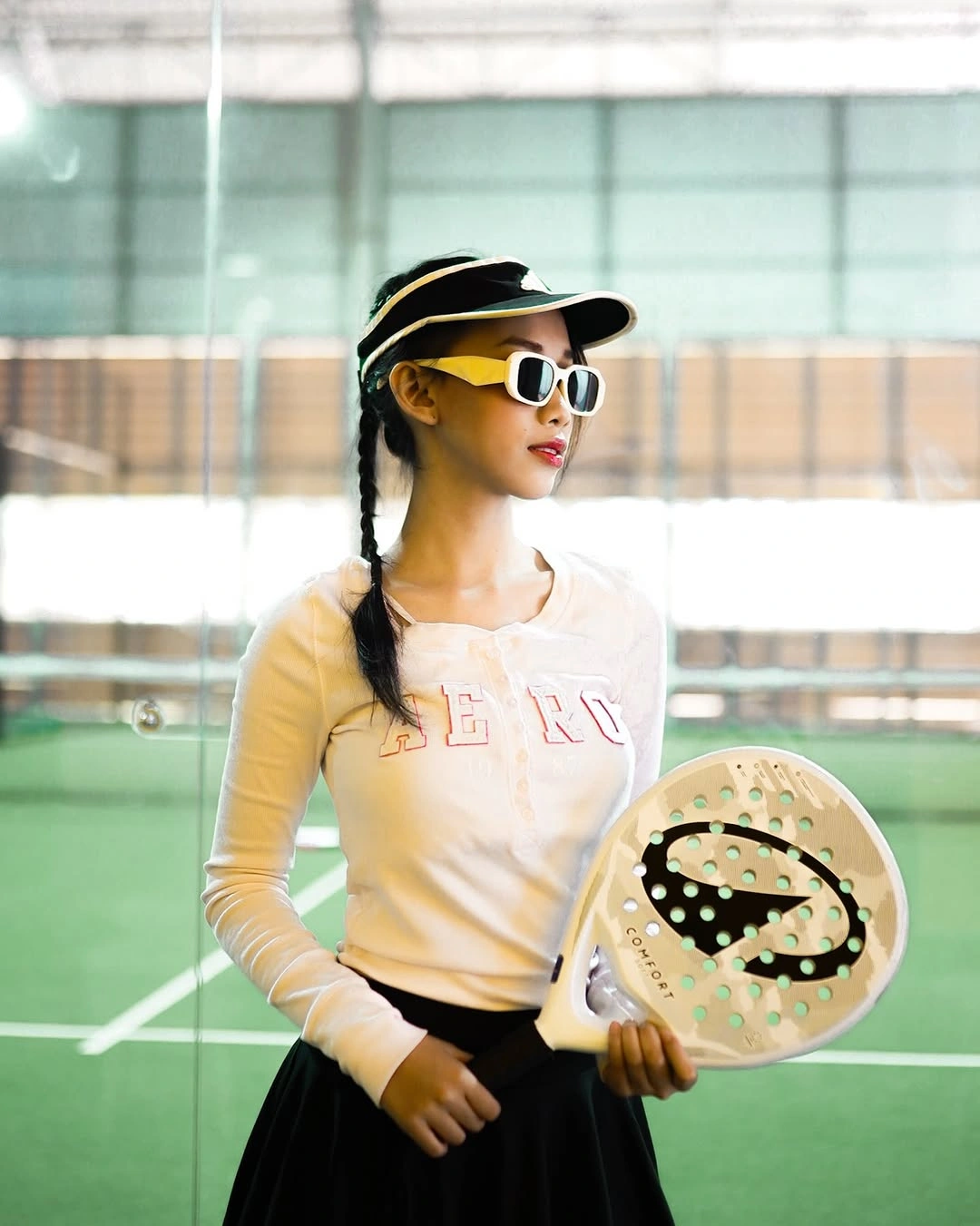 Cream long sleeve athletic top black pleated skirt visor sunglasses and padel racket