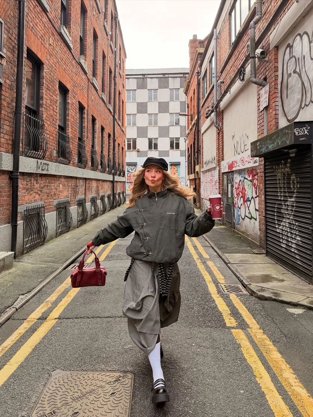 Olive bomber with gray draped trousers, white socks, and burgundy bag